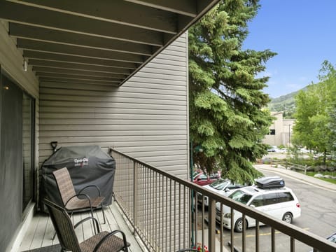 A balcony with two chairs, a barbecue grill, and a view of parked cars and trees. A building and hilly landscape are visible in the background.