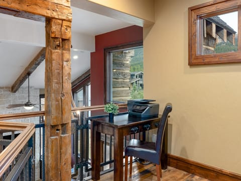 A small office nook with a wooden desk, chair, and printer, situated by a window overlooking a rustic outdoor view. The space is framed by wooden beams and railing.