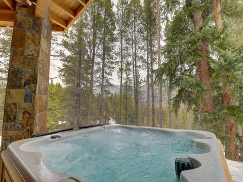 A hot tub under a covered wooden structure, surrounded by tall pine trees with a mountainous landscape in the background.