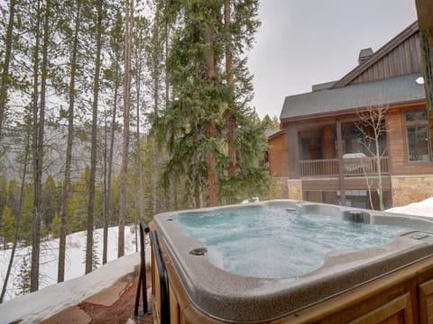 A hot tub on a snowy deck overlooks a wooded area and a rustic cabin. Trees surround the scene, with mountains visible in the background. Steam rises from the bubbling water in the tub.