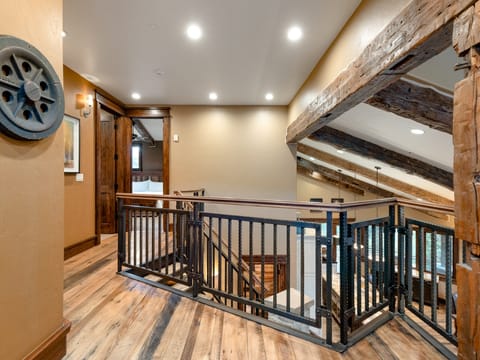 A wooden staircase with metal railing leads to an open hallway with hardwood floors, exposed ceiling beams, and warmly lit walls.