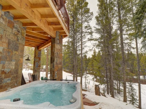 An outdoor hot tub on a snowy day is situated under a wooden deck, with tall pine trees and a forested area in the background.