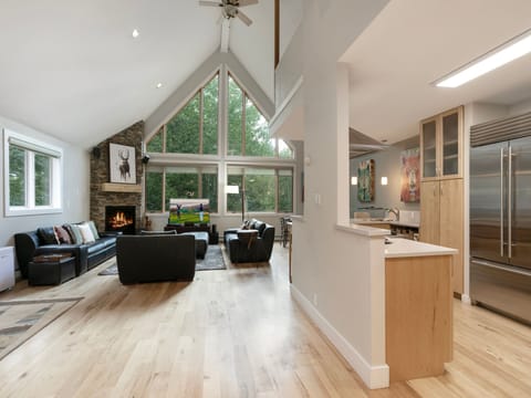 Open-concept living space with hardwood floors, large windows, and a fireplace on the left. A modern kitchen with light wood cabinets is on the right. A wall-mounted TV is on in the background.