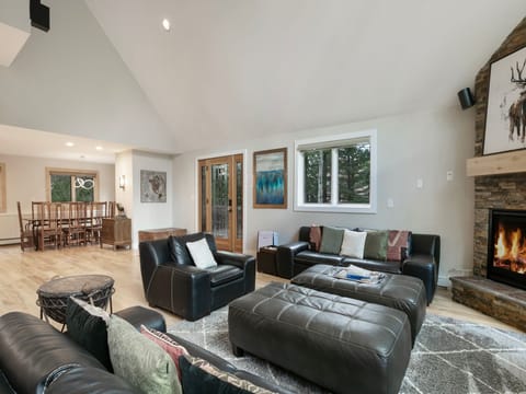 A modern living room features black leather sofas, a large ottoman, a fireplace with a mounted deer head, and a view of the dining area and kitchen in the background.