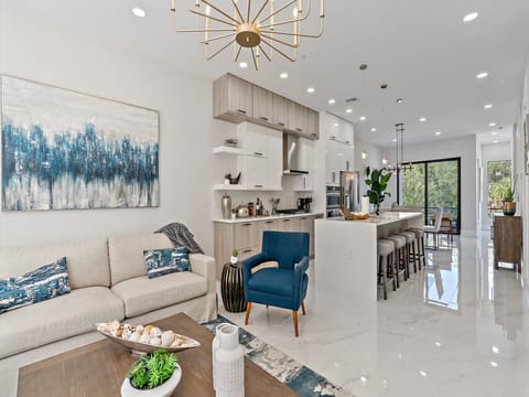 Modern open-concept living room and kitchen with white cabinetry, a beige sofa, blue armchair, island with bar stools, and decorative lighting. A large abstract painting hangs above the sofa.