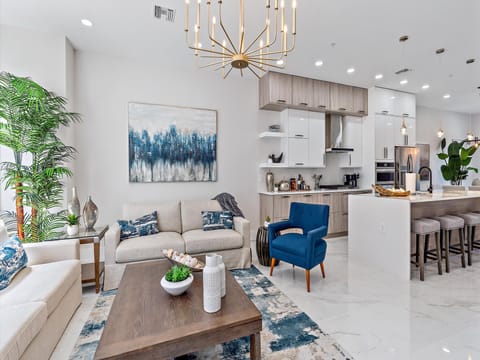 Modern living room and kitchen with white walls, hanging light fixture, blue and beige furniture, a large plant, abstract wall art, and a wooden coffee table with decorative items.