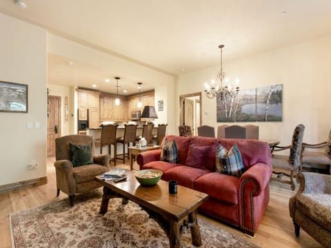 A warmly lit living room with a red sofa, patterned rug, rustic wooden coffee table, and two armchairs. The background features a kitchen with wooden cabinets and a dining area with a chandelier.