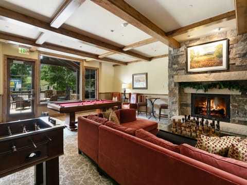 A cozy recreation room features a pool table, foosball table, red sofas, a chess set, and a stone fireplace. Large windows and doors open to an outdoor area surrounded by trees.