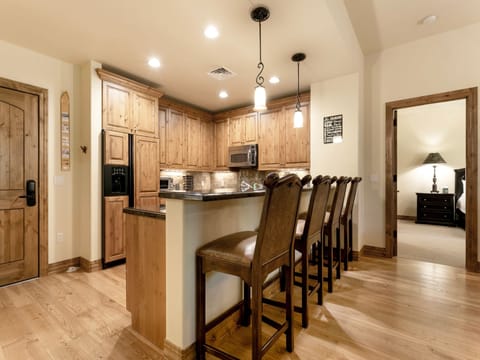 A kitchen with wooden cabinetry, a black countertop, a stainless steel microwave and stove, pendant lights, and four tall brown chairs at the island. An adjacent room with a bed is visible in the background.