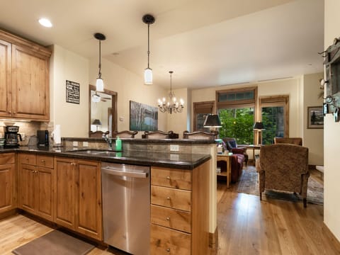 A modern kitchen with wooden cabinetry, stainless steel appliances, and a granite countertop opens into a living area with a chandelier, sofa, and large windows.