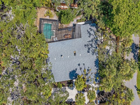 Aerial view of a house surrounded by trees, featuring a screened-in pool area and an outdoor seating space. The roof has two skylights and the property has a mix of green areas and paved walkways.