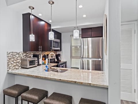 Modern kitchen with a granite countertop bar featuring three stools, stainless steel appliances, and dark brown cabinets. Pendant lights hang above the counter.