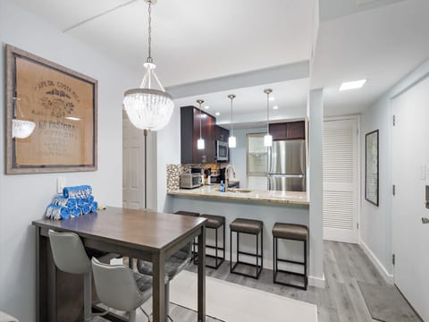 A kitchen with dark wood cabinets, stainless steel appliances, and a beige stone countertop. A dining table with gray chairs is nearby, and a pendant light hangs above. A framed artwork is on the wall.