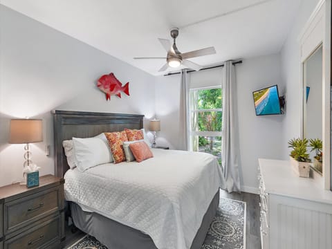 A bedroom featuring a large bed with white bedding, two nightstands with lamps, a mounted fish decoration, a ceiling fan, a wall-mounted TV, a dresser with decor, and a window with white curtains.