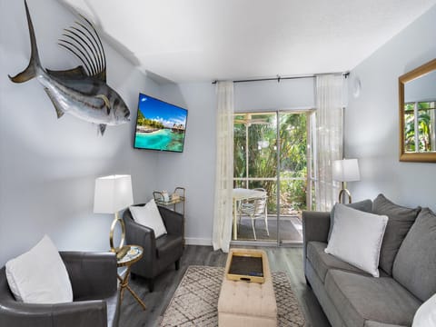 A modern living room features a mounted fish, TV, two chairs, a sofa, a coffee table, and sliding glass doors leading to a patio with outdoor dining furniture and views of lush greenery.