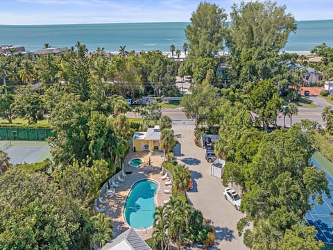 Aerial view of a residential area with a swimming pool, tennis court, trees, and nearby beach with calm blue waters in the background.