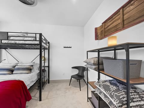 A bedroom with a bunk bed, a shelf holding folded blankets and storage baskets, a chair, and a small lamp. A wooden canoe decoration is mounted on the wall above the shelf.