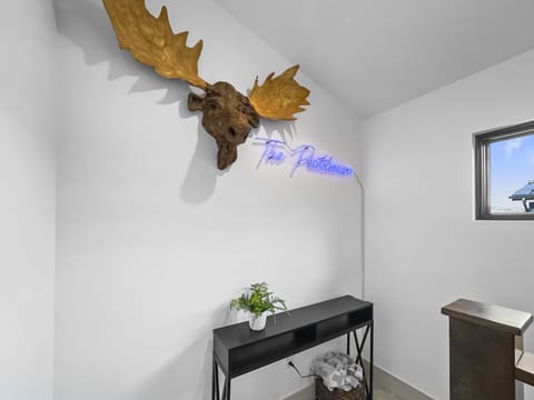 A wall-mounted moose head sculpture above a black console table with a potted plant and a wicker basket beneath. A neon sign reading "The Penthouse" is illuminated beside the sculpture.