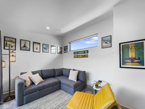 A modern living room featuring a grey sectional sofa, a yellow chair, wall art, and a tall floor lamp. The walls are decorated with vintage posters and a "High Country Rd Iowa Hawkeye Dr" sign.