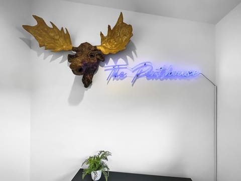 A wall-mounted golden moose head sculpture and a neon sign that reads "The Penthouse" are displayed above a small table with a potted plant.