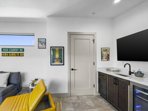 A small room with a TV, framed pictures, a yellow chair, a gray couch, a cabinet with a countertop, and decor signs on the wall. A closed door is in the center of the image.