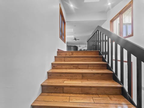 A wooden staircase with a black railing leads up to a bright, open hallway featuring windows and a ceiling fan.