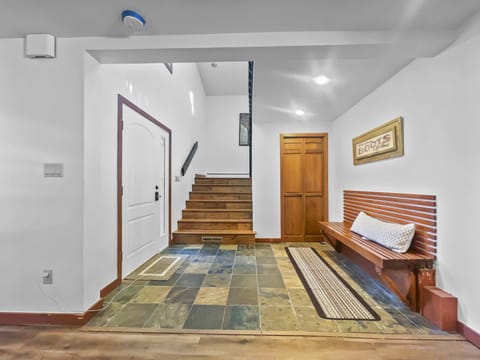 A foyer with slate tile flooring, a white front door, wooden stairs, a wooden bench with a cushion, and a framed "Boots" sign on the wall.