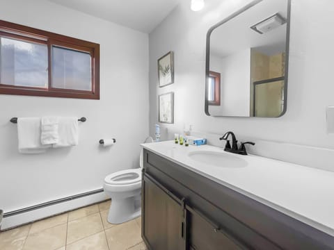 Modern bathroom with a white countertop sink, rectangular mirror, toilet, towel rack, and two framed pictures on the wall. The room has beige tile flooring and a window with wooden trim.