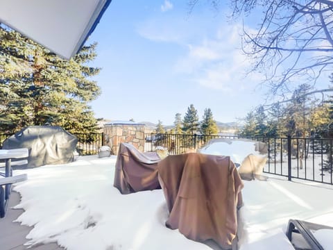 Snow-covered outdoor patio with furniture covered in protective tarps, surrounded by trees and overlooking a distant mountain range under a clear sky.