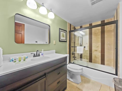 Modern bathroom with green walls, white ceiling, and recessed lighting. Features a vanity with a rectangular mirror, a toilet, and a glass-enclosed shower-tub combination with striped tile accent.