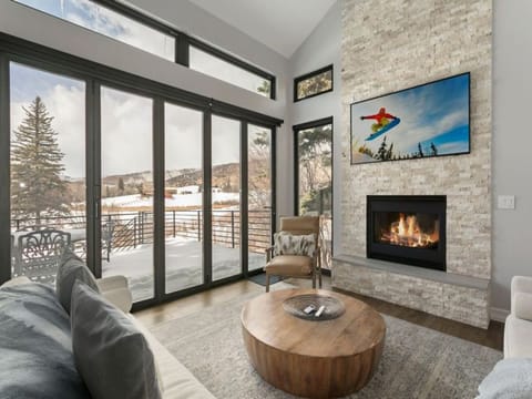 A modern living room with large glass windows, a fireplace, a round coffee table, and a flat-screen TV displaying a snowboarder. Snowy landscape visible outside.