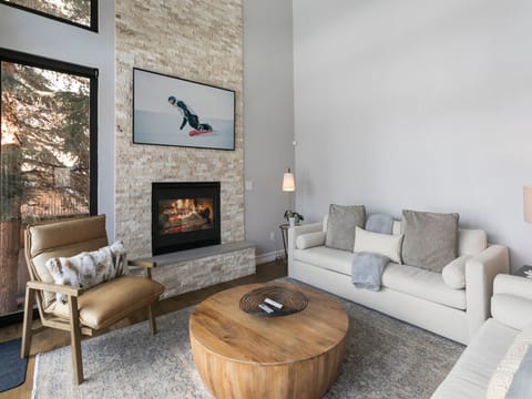 A modern living room features a fireplace beneath a photo of a snowboarder, a white couch, a beige armchair, a wooden coffee table, and floor-to-ceiling windows with a view of trees.