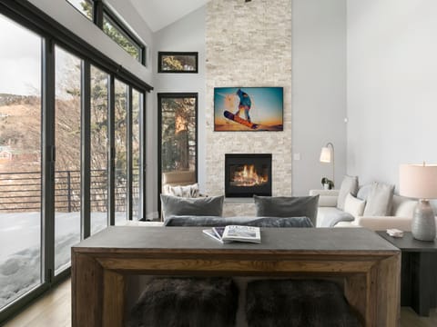 Modern living room with large windows, a stone fireplace, a wooden table with a lamp, and a sofa. A snowboarder picture hangs above the fireplace. Outside view of trees and snow through the windows.