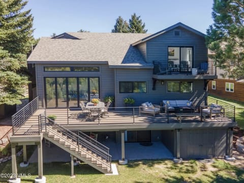 Two-story gray house with a large wooden deck surrounded by trees, featuring outdoor furniture on the deck, large glass windows, and a staircase leading to the backyard.