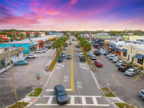 Aerial view of a busy street lined with shops, cars parked on both sides, and a vibrant purple and pink sunset in the background.