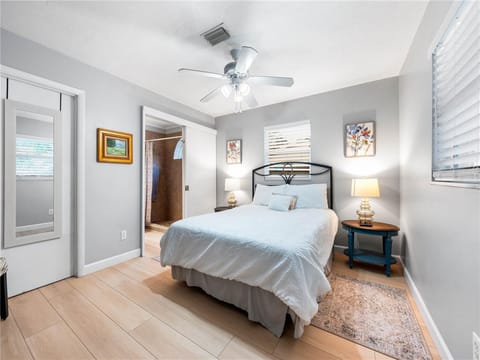 A bedroom with a queen bed, two nightstands with lamps, an overhead fan, a small rug, two paintings on the wall, and a view into an adjacent bathroom. The room has light wood flooring and white walls.