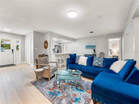 A cozy living room with a blue sectional sofa, a glass coffee table, and a colorful rug. There is a small dining area and a kitchen in the background, with a doorway leading to a bedroom.