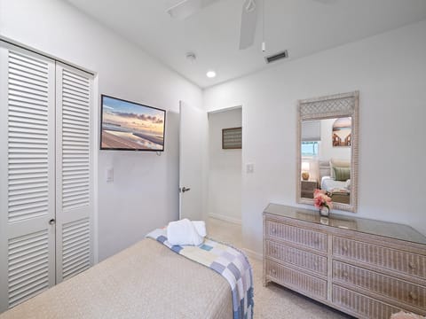 A clean, neatly organized bedroom with a bed, dresser, wall-mounted TV, and a mirror. The room features white walls, a ceiling fan, and a window with a view of another room.