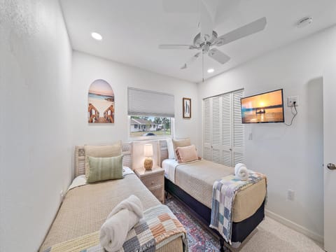 A bedroom with two single beds, each with a rolled towel, a nightstand with a lamp, a wall-mounted TV, and a ceiling fan. A window with blinds and a painting are on the walls.