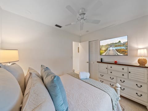 A neatly arranged bedroom with a large bed, white bedding, and blue accent pillows. A ceiling fan is above, a dresser with a TV and lamp on top is against the wall, and a doorway is visible.