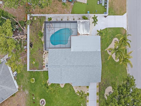 Aerial view of a house with a screened swimming pool and surrounding yard, adjacent to a driveway and street. The property features trees and landscaped areas.