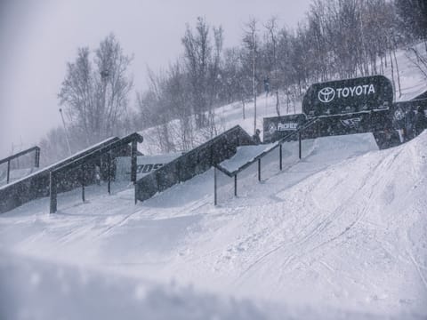 Snow-covered winter sports park with various rails and ramps, featuring Toyota advertising signage in the background. Trees and clouded sky visible.