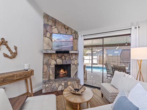 A cozy living room with a stone fireplace, wall-mounted TV, and beige seating. Sliding glass doors lead to an outdoor pool area with patio furniture. A wood table and decorative anchor are on the left.