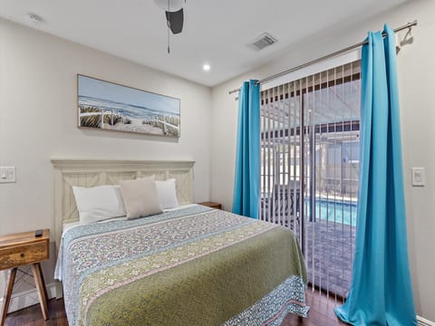 A bedroom with a large bed featuring a colorful quilt, two nightstands, and blue curtains covering sliding glass doors that open to a view of a backyard pool. A landscape painting hangs on the wall above the bed.