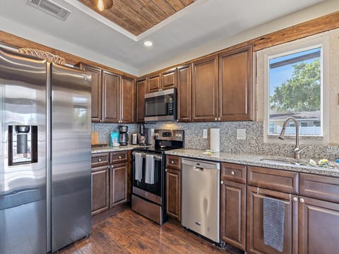 A modern kitchen with dark wooden cabinets, stainless steel appliances including a refrigerator, oven, microwave, and dishwasher, a granite countertop, and a window above the sink.