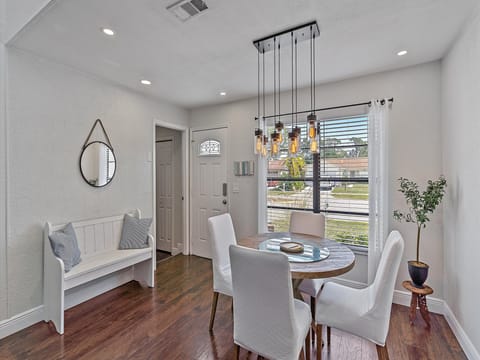 A dining room with a round table surrounded by four white chairs, a bench with pillows, a hanging light fixture, a potted plant, a wall mirror, and a window with blinds and a curtain.