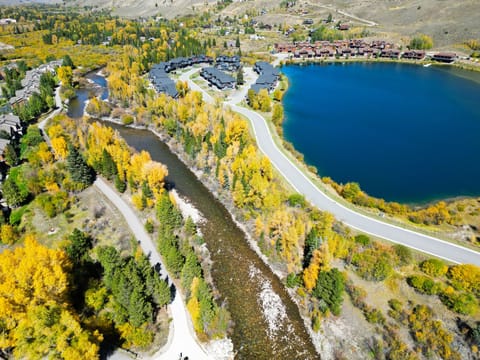 Aerial view of a scenic landscape featuring a clear blue lake, flowing river, surrounding roads, and buildings, all amidst vibrant autumn foliage.