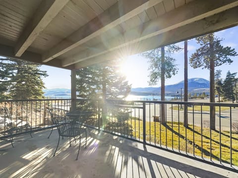 A covered patio with outdoor furniture overlooks a scenic view featuring trees, a road, and a body of water, with the sun shining brightly in the background.