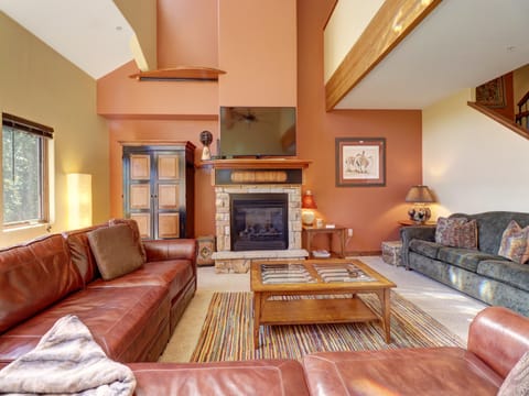 A living room with brown leather sofas, a striped rug, a fireplace with a mounted TV, a wooden coffee table, and an upper balcony. The walls are painted in shades of cream and beige.