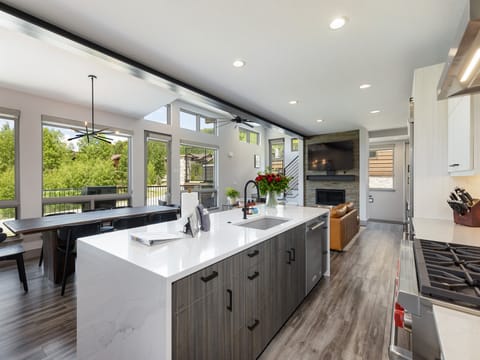 A modern kitchen with a large island, white countertops, stainless steel appliances, wood cabinetry, and a view of the living area with a fireplace and large windows looking out to greenery.
.